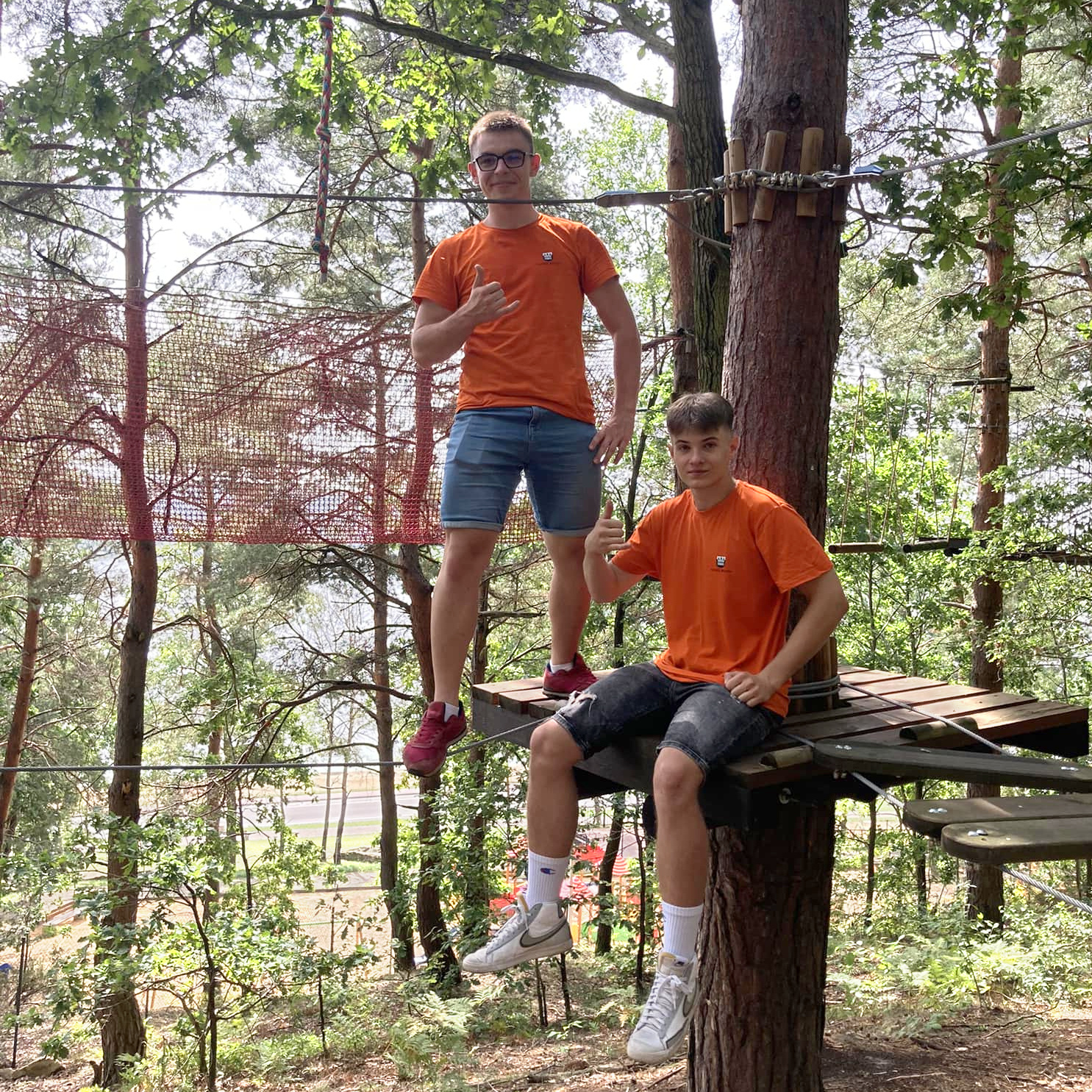 Dwaj chłopcy w parku linowym obrani w pomarańczowe koszulki. Są to operatorzy parku linowego.