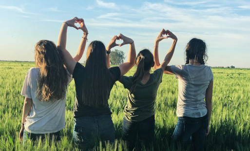 Grupa młodzieży widziana z tułu tworzy z dłoni kształty serc. Młodzi siedzą na trawie.